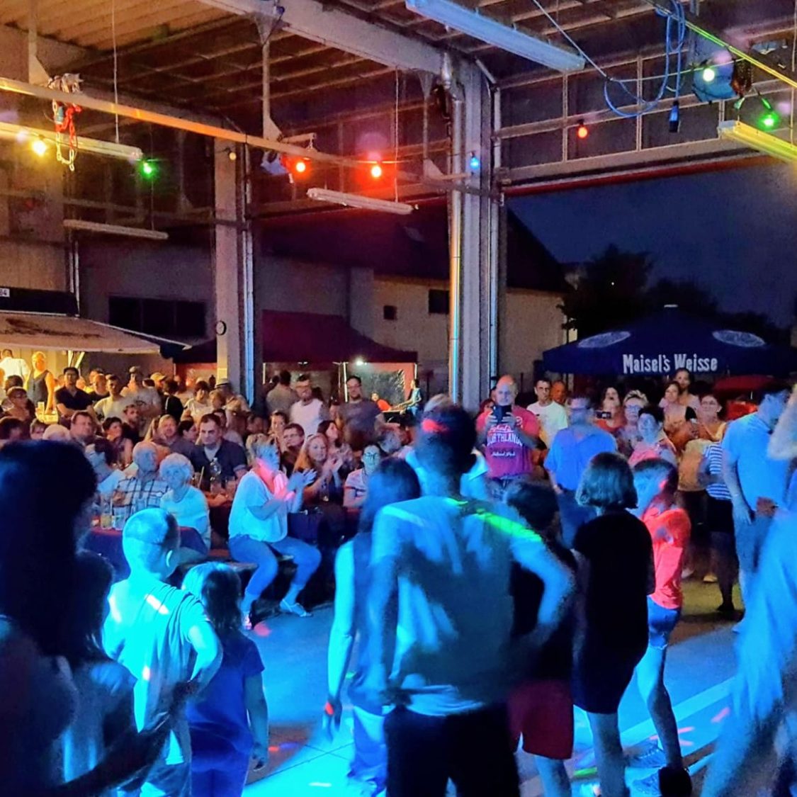 Bunte Party mit Menschen tanzend und feiernd in einem Feuerwehgerätehaus bei Nacht.