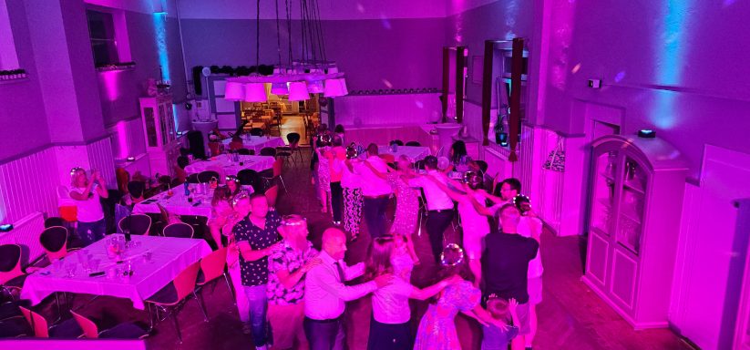 Menschen tanzen in einem Raum mit bunten Lichtern und einer festlichen Atmosphäre.
