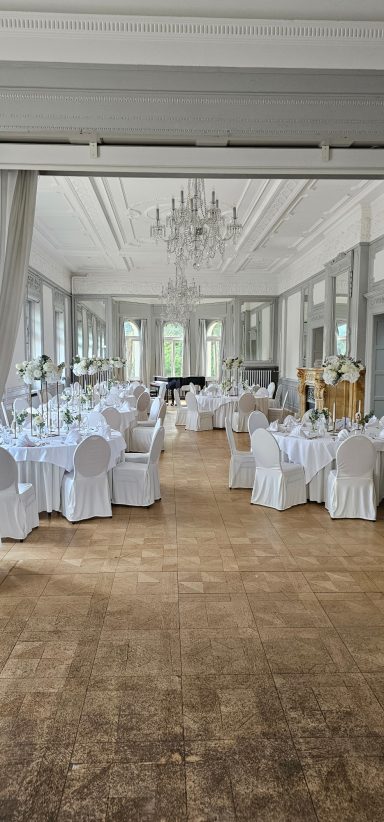 Eleganter Festsaal mit runden Tischen, weißen Stühlen und Kronleuchter.